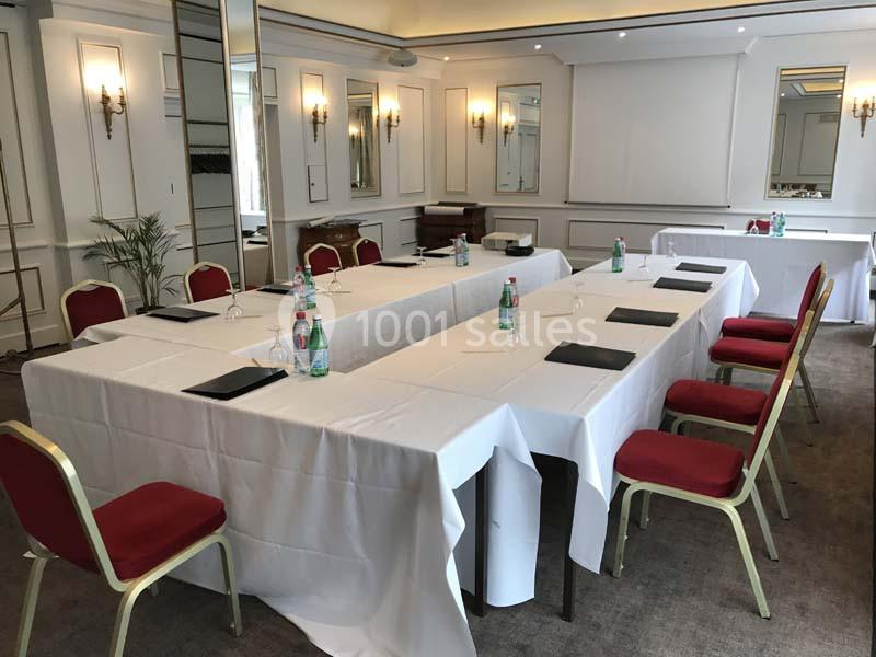 Salle de réunion lumineuse avec tables disposées en U, nappées de blanc, chaises rouges et bouteilles d'eau.