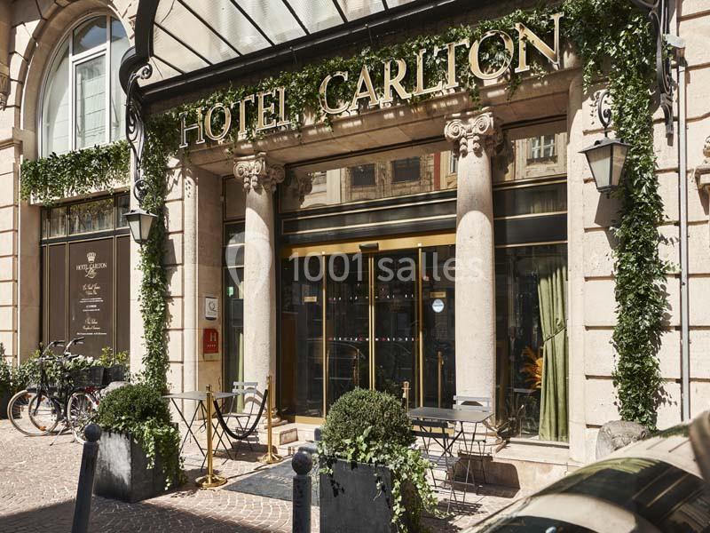 Façade d'un hôtel avec enseigne ’Hôtel Carlton’, terrasse aménagée et végétation décorative.