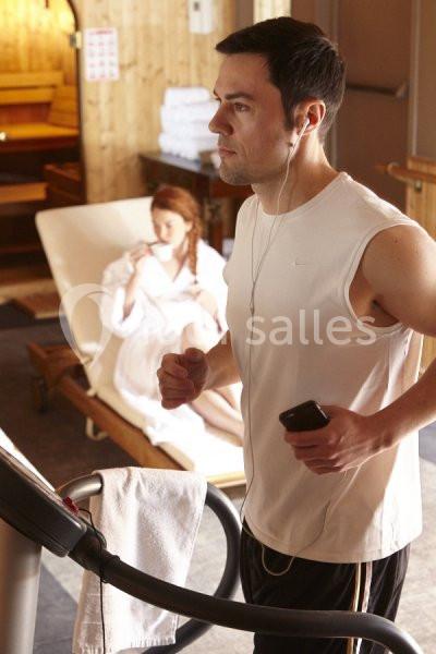 Un homme court sur un tapis de course dans un spa, une femme en peignoir est assise en arrière-plan.