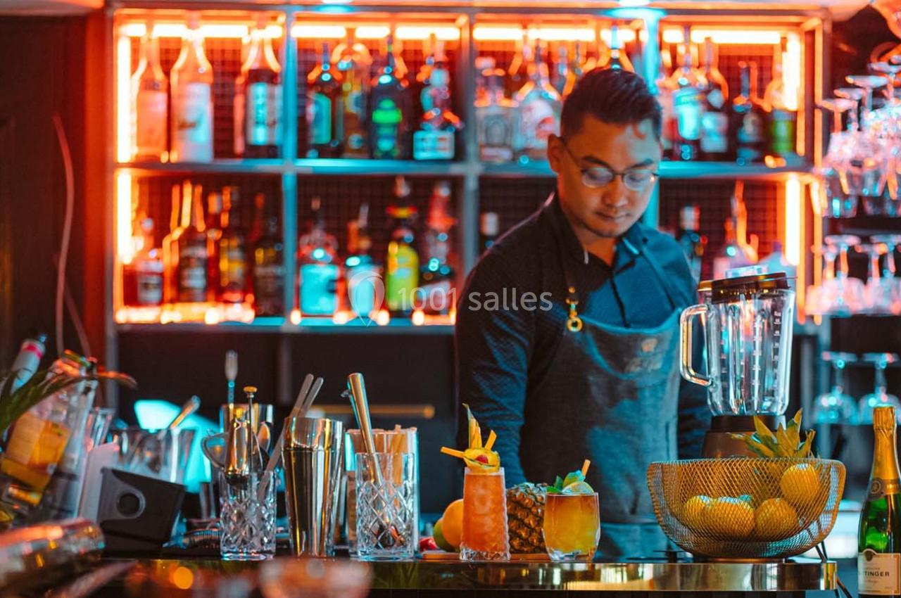 Un barman prépare des cocktails dans un bar éclairé par des lumières colorées, avec des bouteilles en arrière-plan.