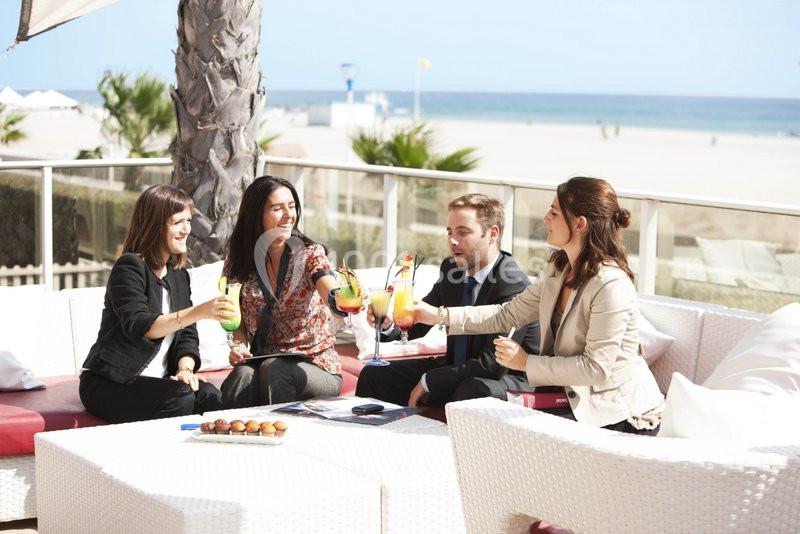 Quatre personnes assises en terrasse, trinquant avec des cocktails colorés, près d'une plage ensoleillée.