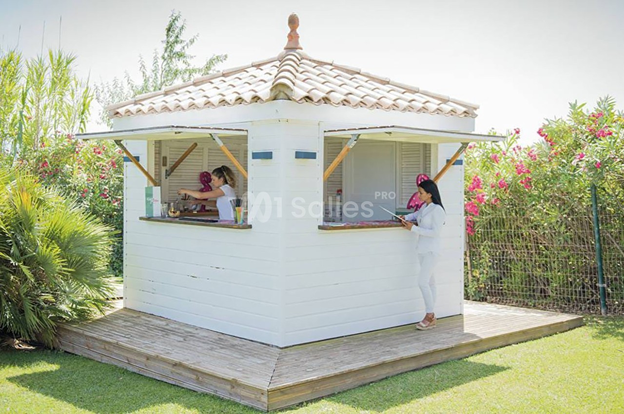 Kiosque en bois blanc avec toit en tuiles, entouré de végétation, utilisé comme espace de service extérieur.