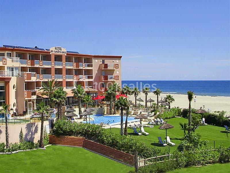 Vue d'un hôtel en bord de mer avec piscine, palmiers, pelouse aménagée et accès direct à une plage de sable.