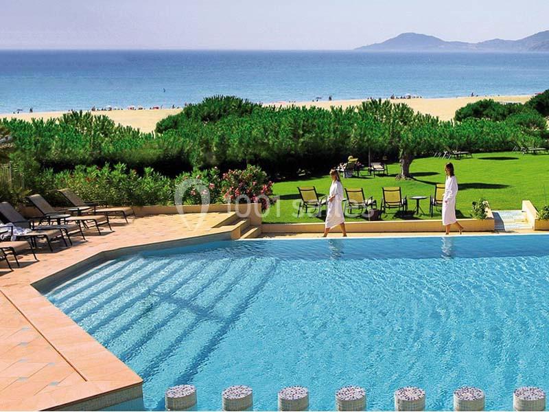 Vue sur une piscine extérieure entourée de chaises longues, avec pelouse, plage et mer en arrière-plan.