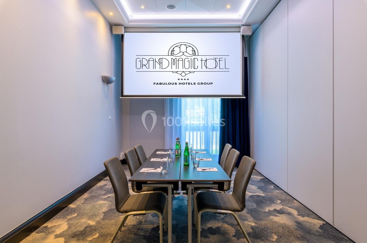 Salle de réunion avec table rectangulaire, chaises, bouteilles d'eau et écran affichant ’Grand Magic Hotel’.