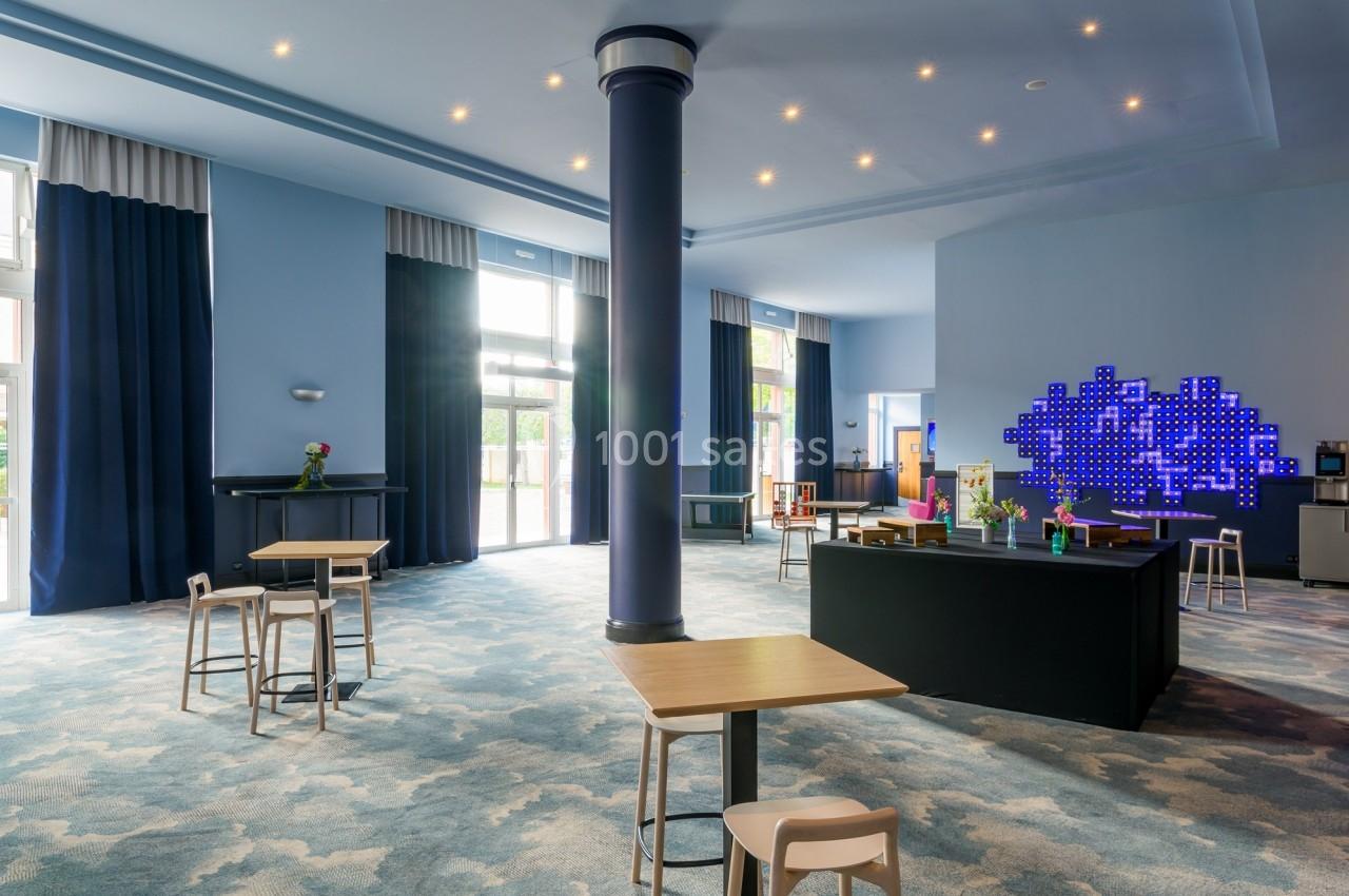 Salle lumineuse avec tables en bois, colonnes bleues, moquette à motifs et mur décoré de lumières LED bleues.