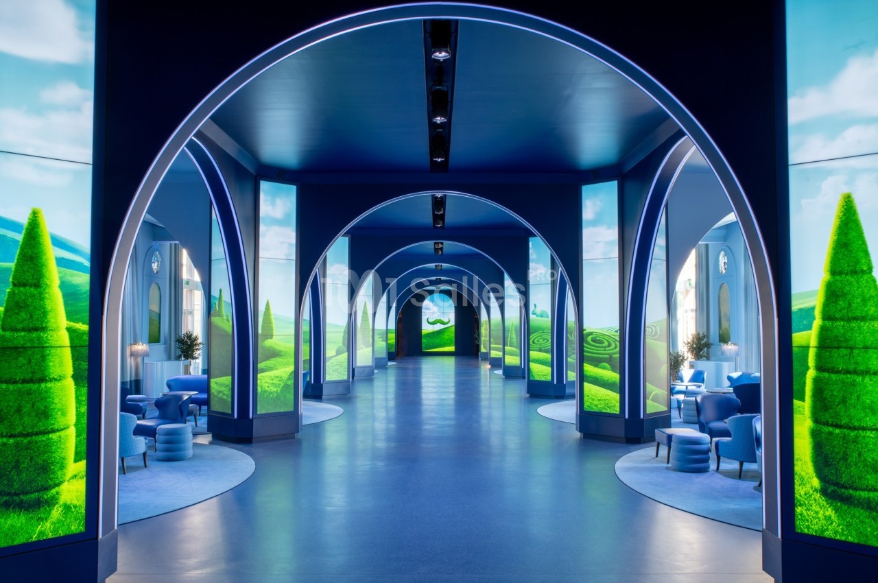 Couloir lumineux avec arches bleues, écrans affichant des paysages verdoyants et mobilier moderne.