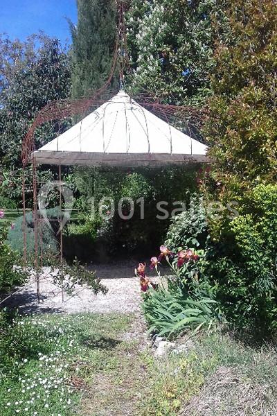 Structure métallique avec un toit blanc en forme de chapiteau, entourée de végétation et de fleurs dans un jardin.