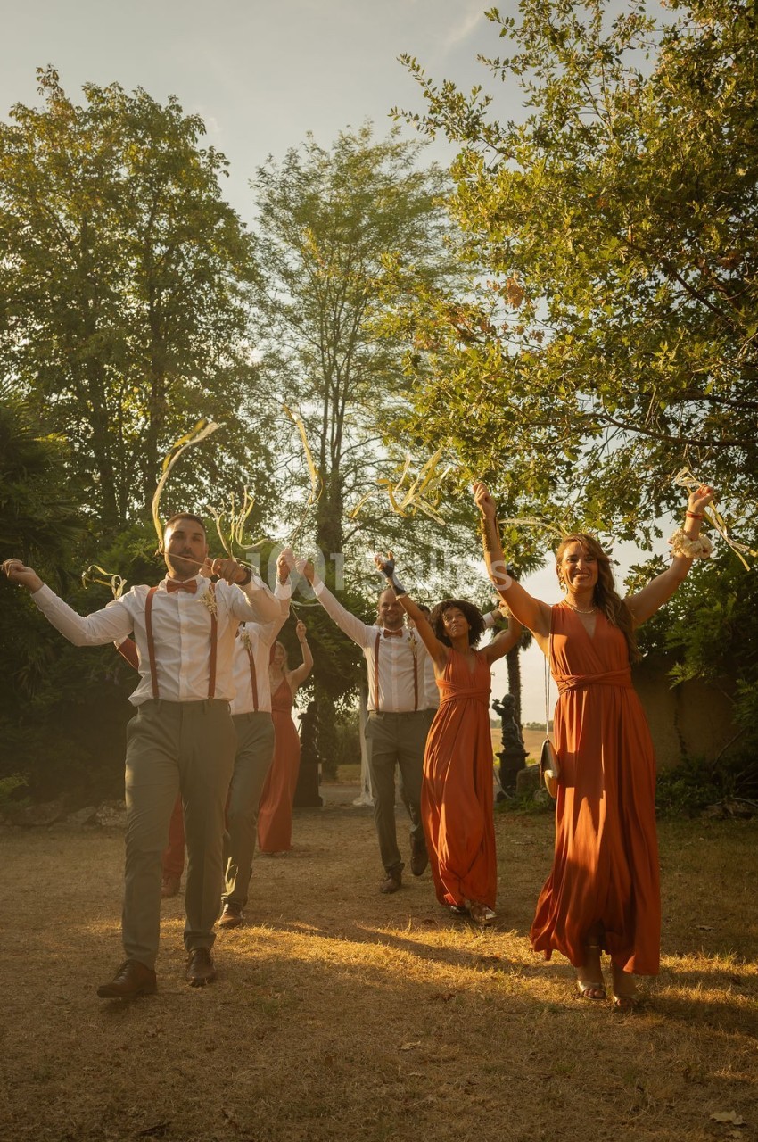 Un groupe de personnes en tenue élégante marche joyeusement dans un jardin ensoleillé, tenant des rubans.