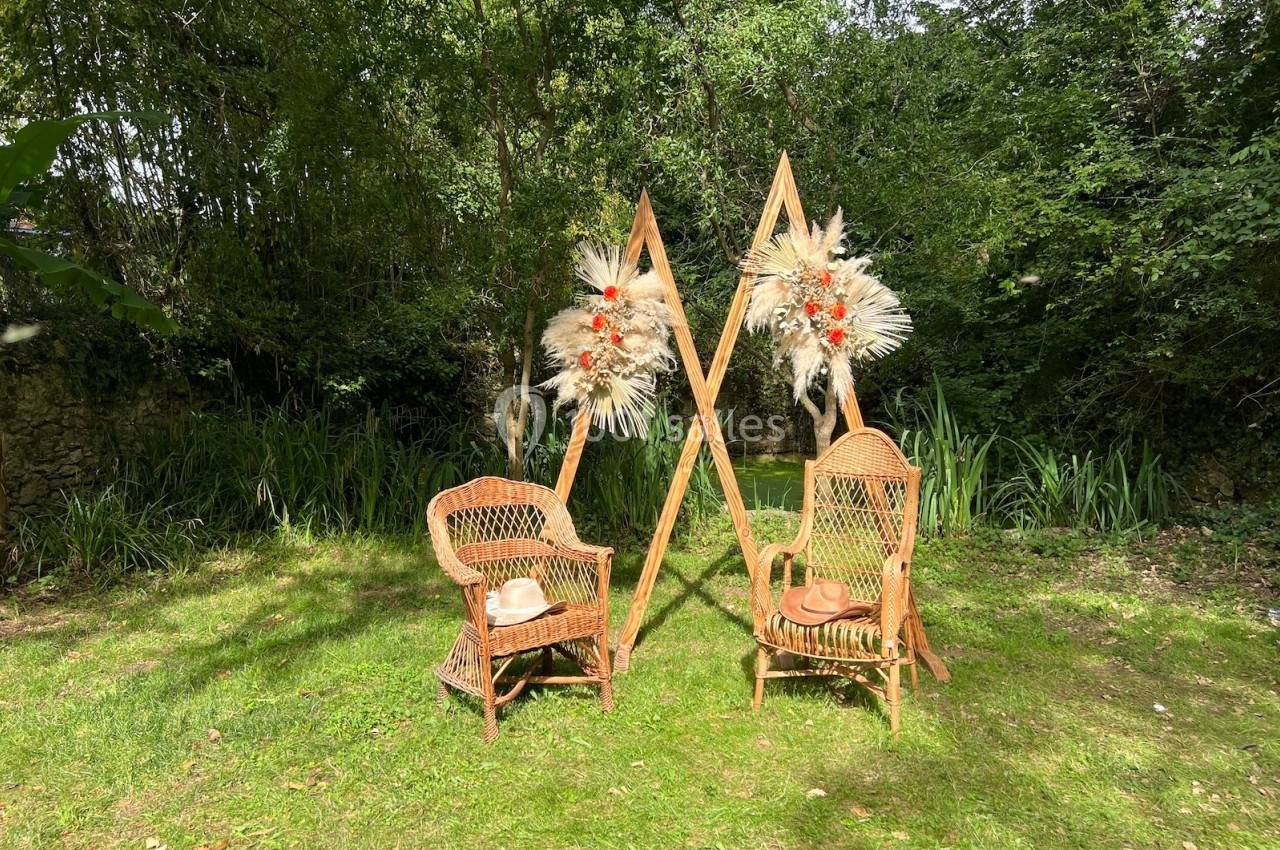 Deux fauteuils en rotin disposés sur une pelouse, encadrés par des structures décorées de fleurs séchées, en extérieur boisé.