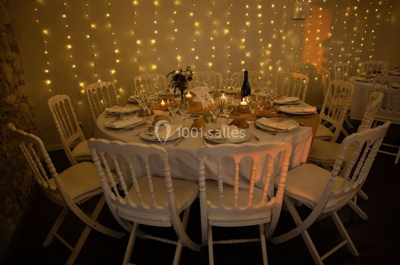 Table ronde dressée pour un dîner, entourée de chaises blanches, avec guirlandes lumineuses en arrière-plan.