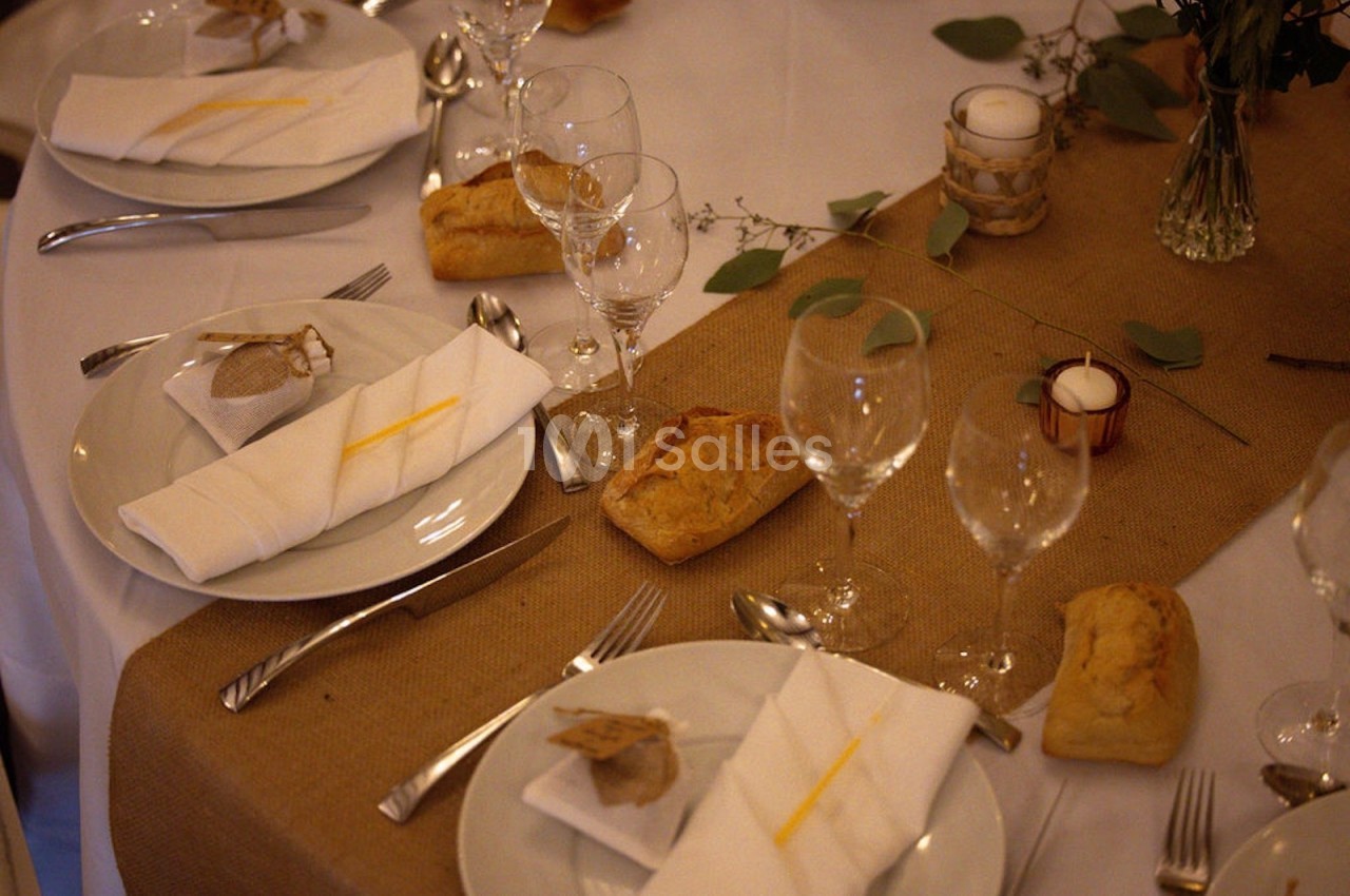 Table dressée avec nappes blanches, chemins de table en jute, verres, couverts, serviettes pliées et petits pains.