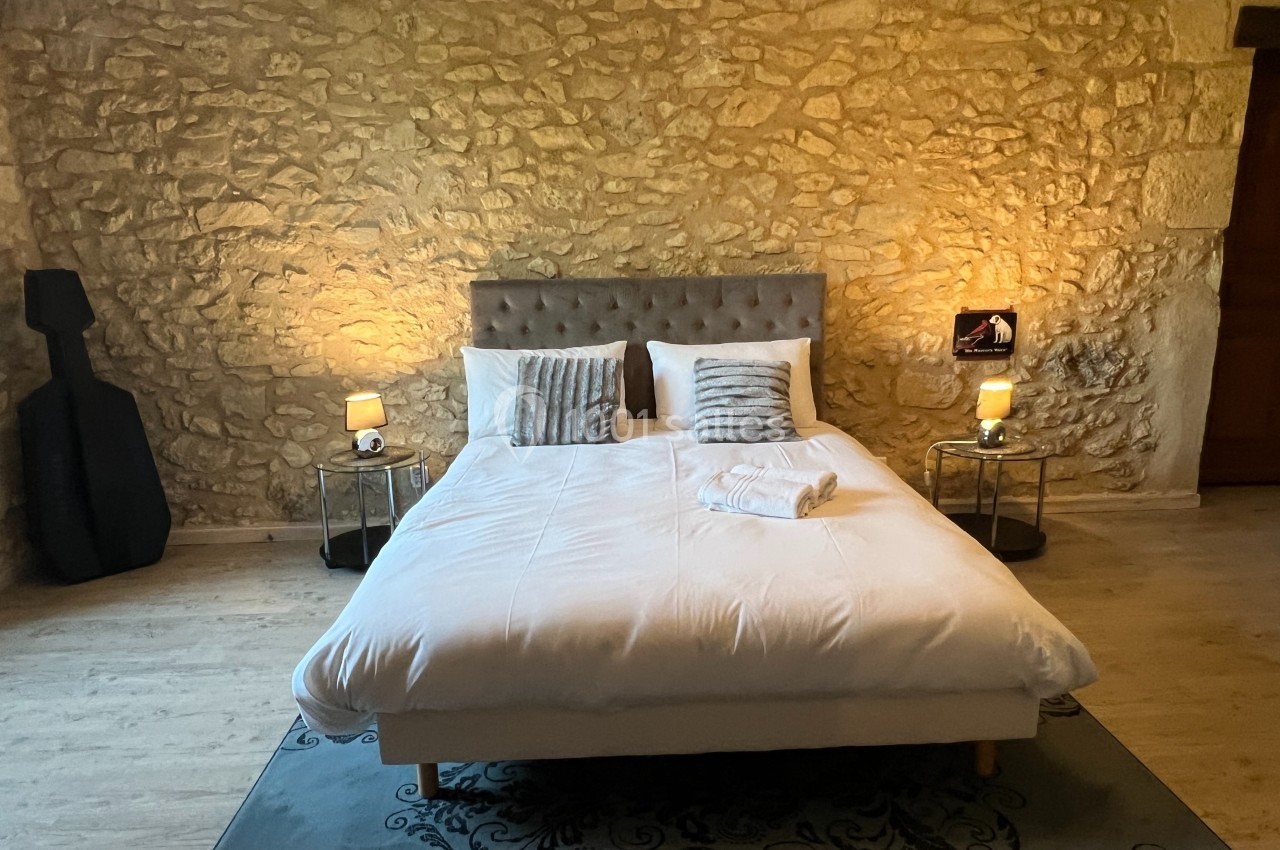 Lit double avec tête de lit capitonnée, draps blancs, coussins décoratifs, éclairage doux et mur en pierre apparente.