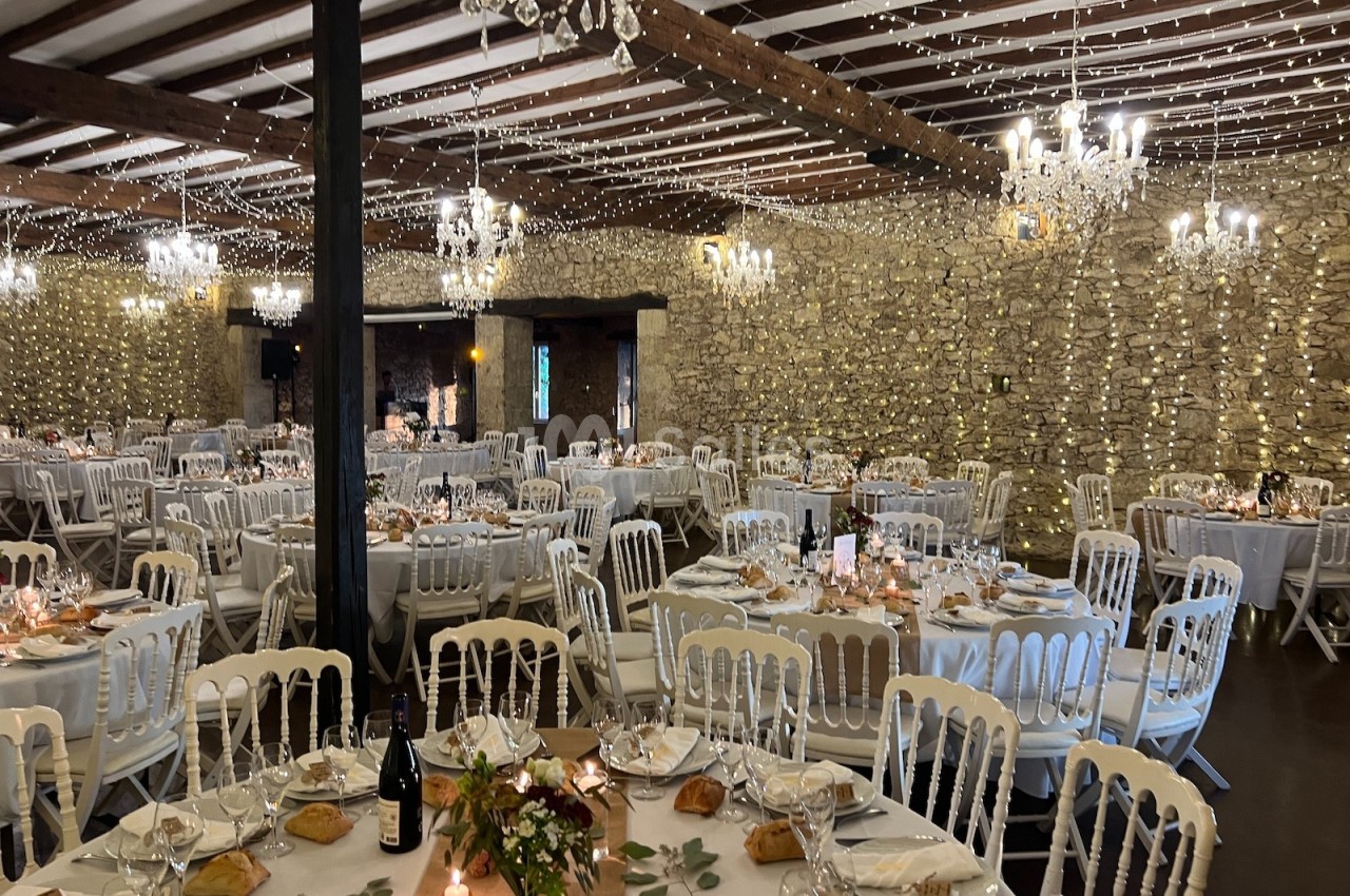 Salle de réception décorée pour un événement, avec tables dressées, guirlandes lumineuses et murs en pierre apparente.