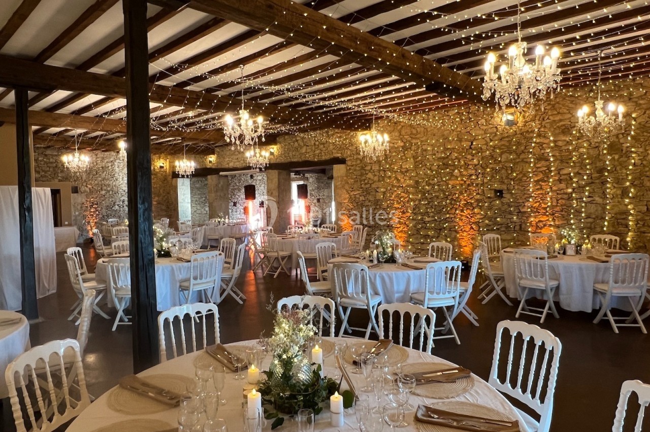 Salle de réception en pierre avec tables rondes dressées, chaises blanches, guirlandes lumineuses et lustres suspendus.