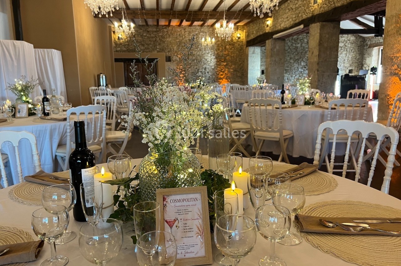 Salle de réception décorée avec des tables rondes, nappes beiges, chandelles, fleurs blanches et chaises blanches.