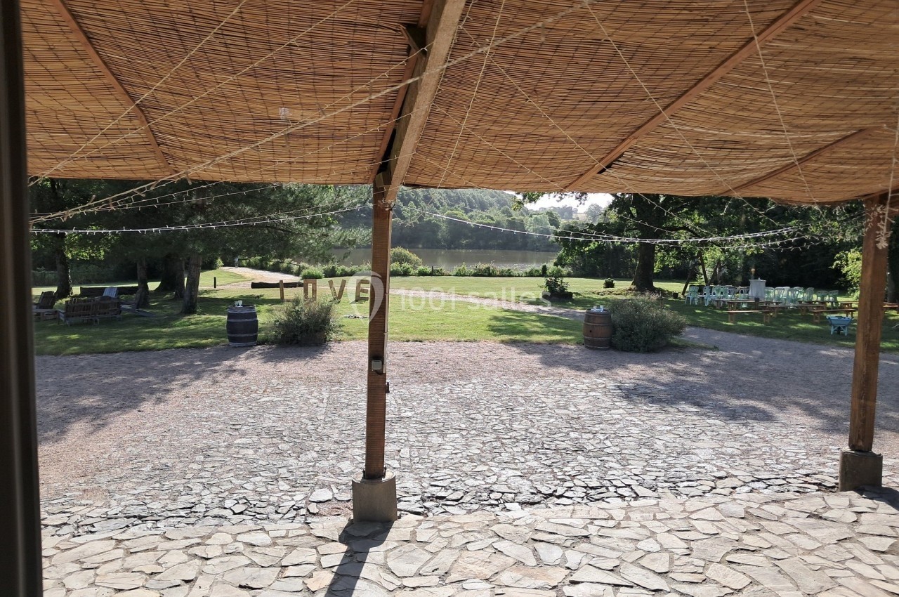Vue d'une terrasse couverte en bois donnant sur un jardin avec pelouse, arbres et rivière en arrière-plan.