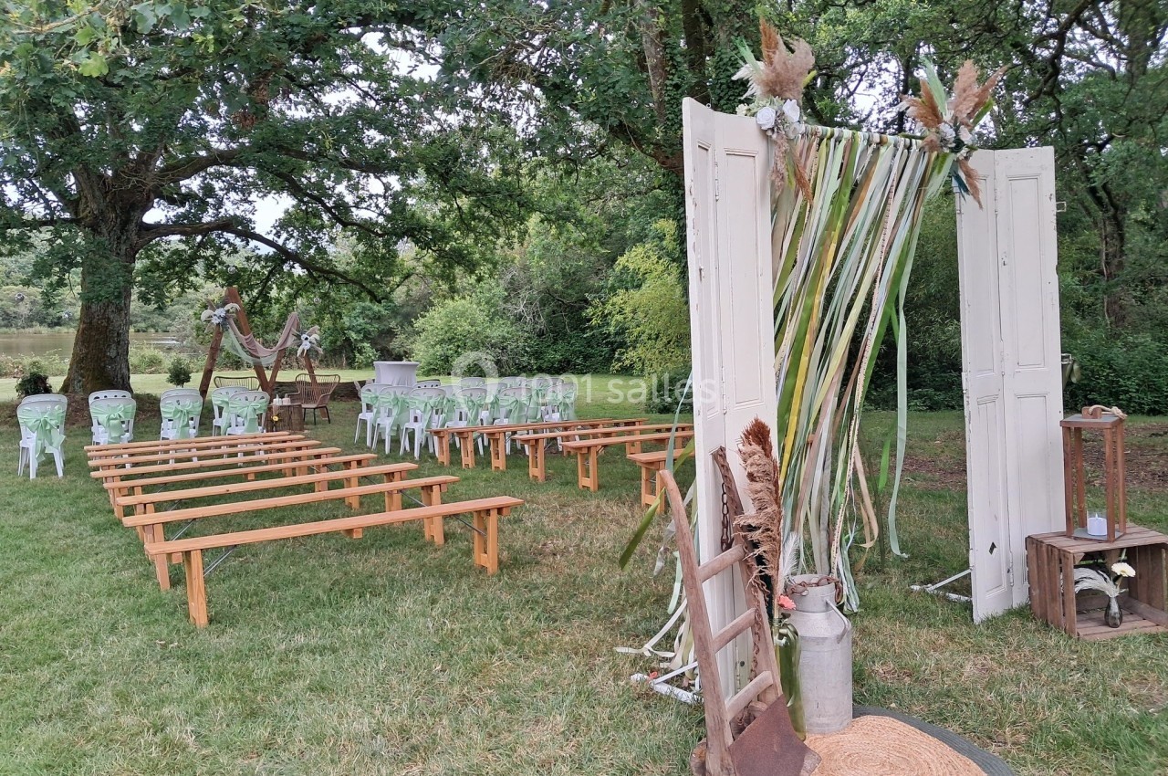 Décoration champêtre pour une cérémonie en plein air avec arche florale, bancs en bois et chaises blanches sur pelouse.