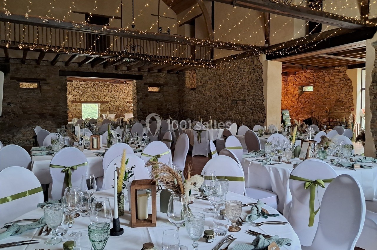 Salle de réception décorée pour un mariage, avec des tables rondes dressées, chaises blanches et guirlandes lumineuses…