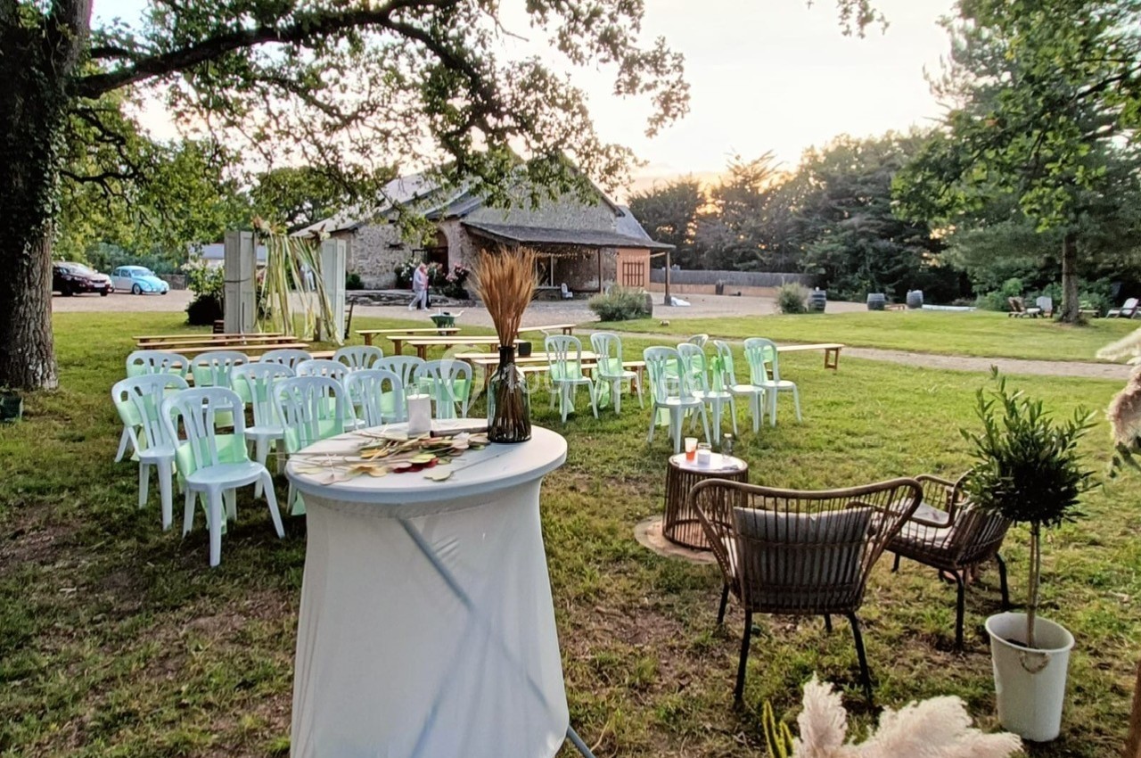 Chaises blanches disposées en extérieur sous des arbres, avec une table décorée et des fauteuils en rotin au premier plan.