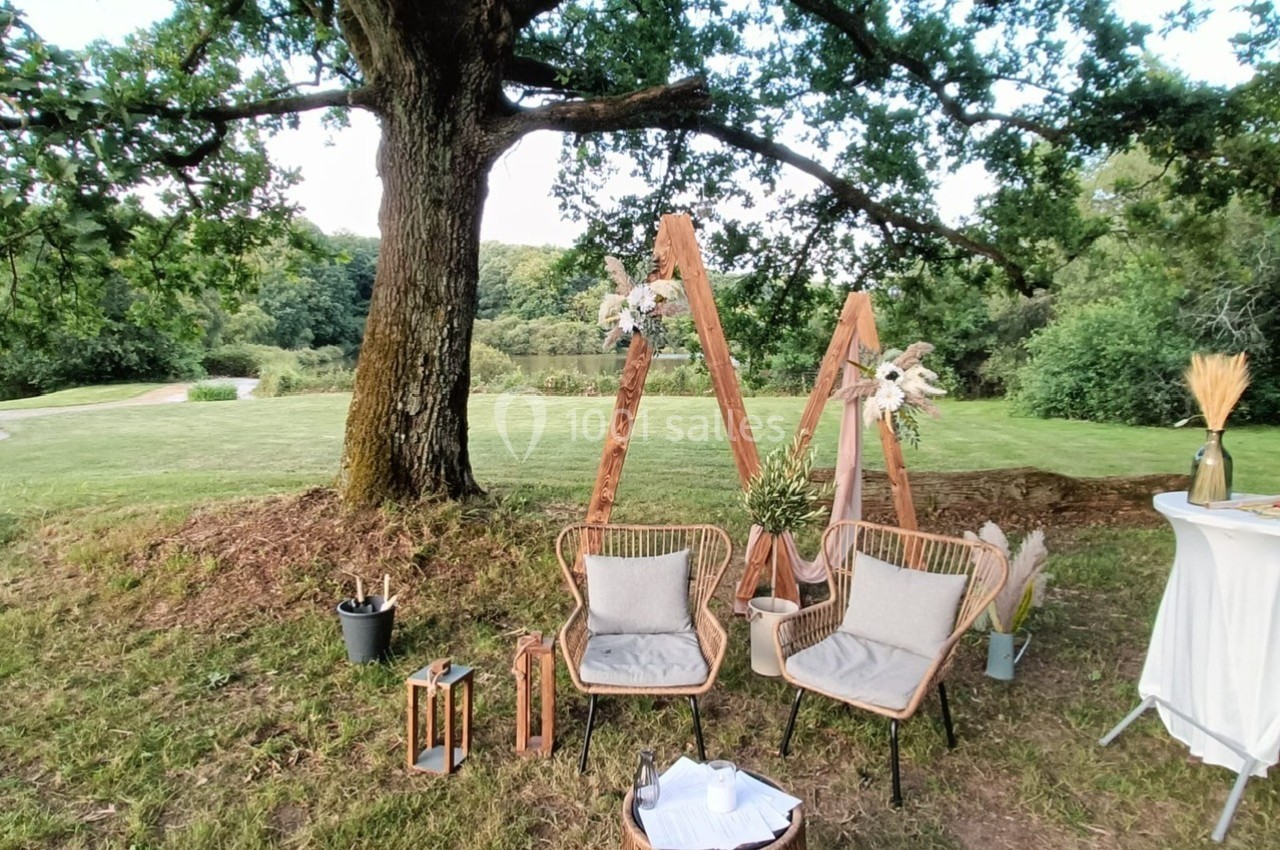 Deux fauteuils en rotin et des décorations boisées disposés sous un grand arbre dans un cadre naturel verdoyant.