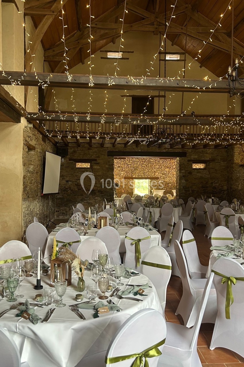 Salle de réception décorée pour un événement, avec tables dressées, chaises blanches et guirlandes lumineuses suspendues.