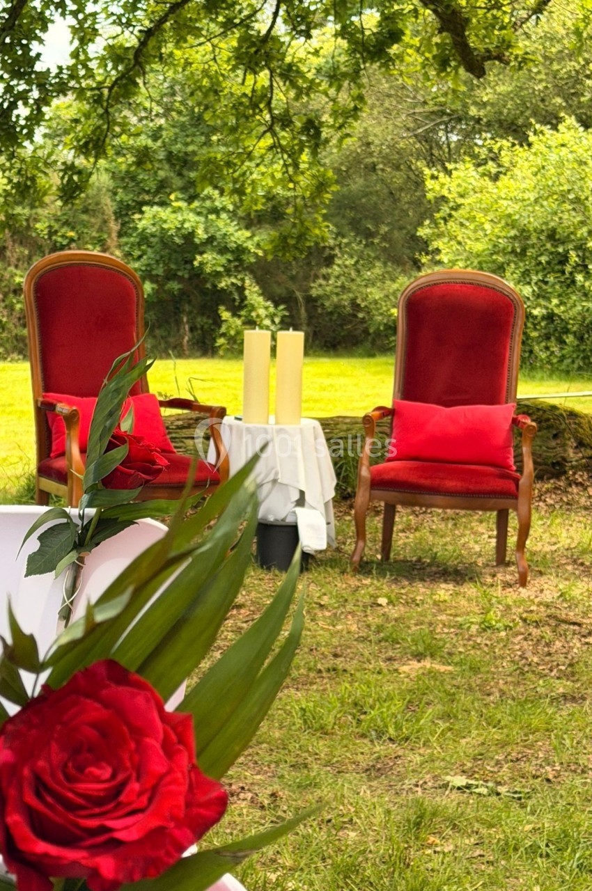 Deux fauteuils rouges disposés sur une pelouse, entourés de verdure, avec des bougies sur une table blanche.