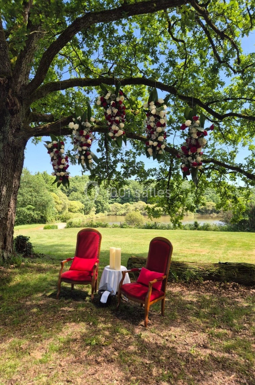 Deux fauteuils rouges sous un arbre décoré de fleurs suspendues, avec une petite table et des bougies, dans un cadre…