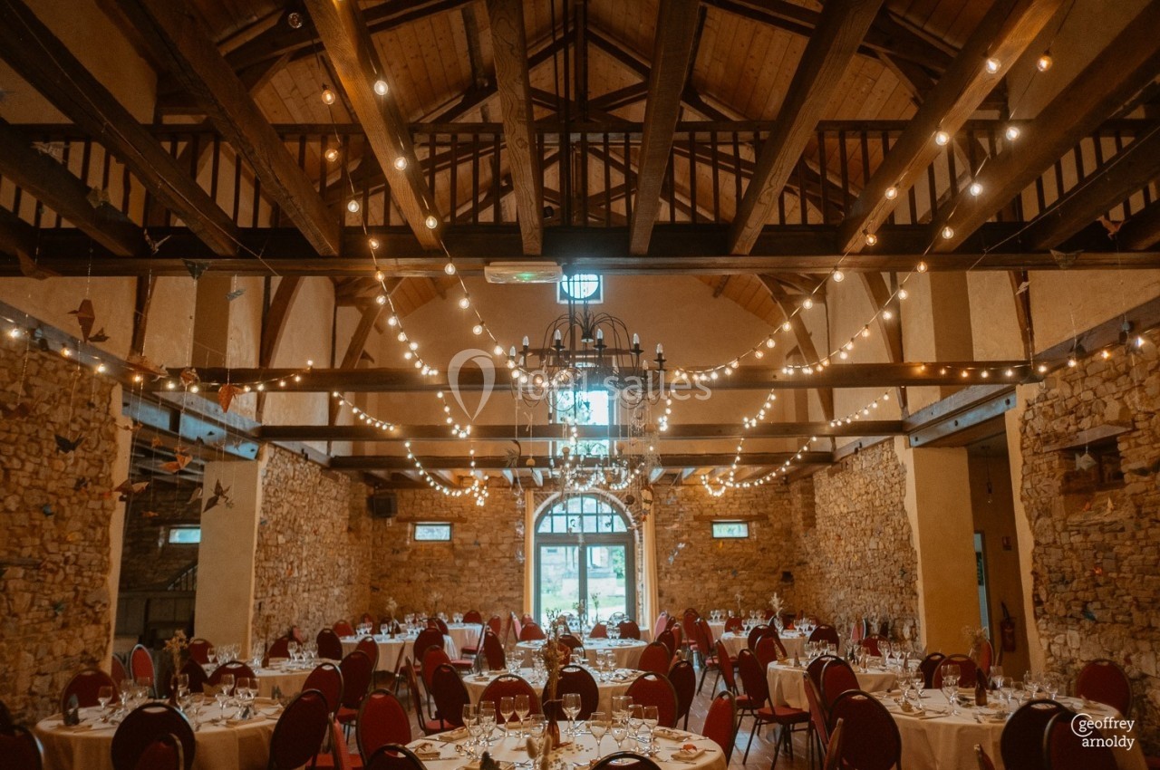 Salle de réception en pierre avec poutres apparentes, tables dressées et guirlandes lumineuses suspendues.
