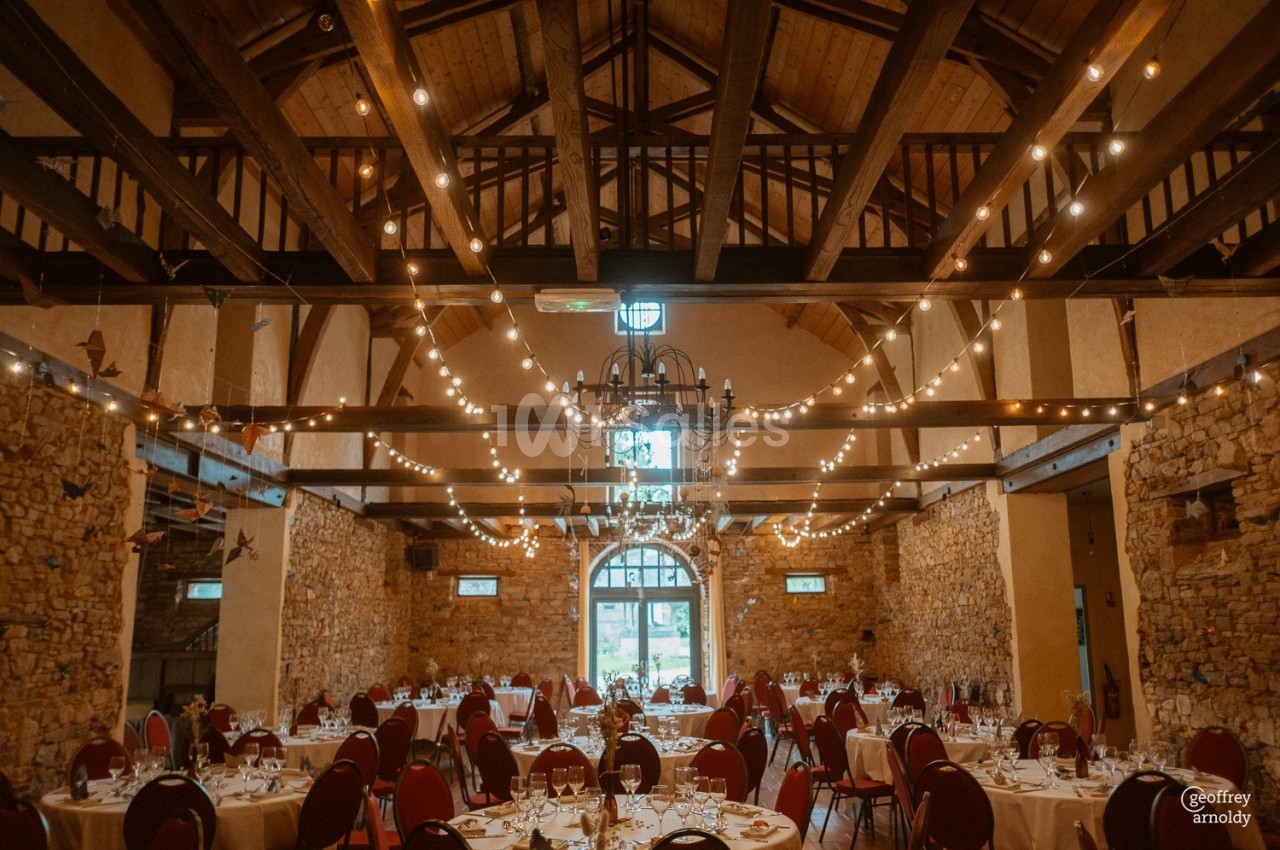 Salle de réception en pierre avec poutres apparentes, tables dressées et guirlandes lumineuses suspendues.