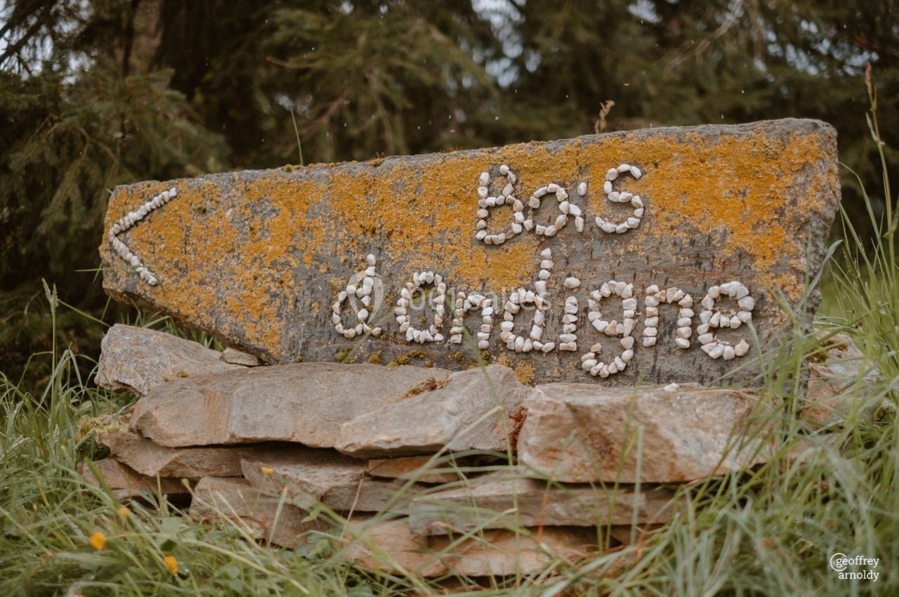 Panneau en pierre recouvert de mousse avec l'inscription ’bas d'ardigne’ formée de petits galets, entouré d'herbe.