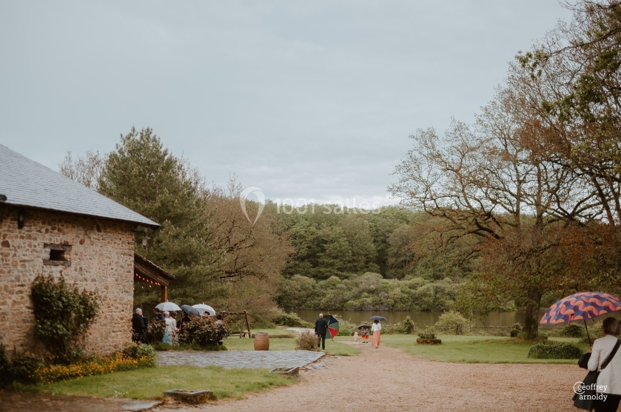 Personnes avec des parapluies marchant près d'un bâtiment en pierre dans un cadre naturel avec arbres et étang.