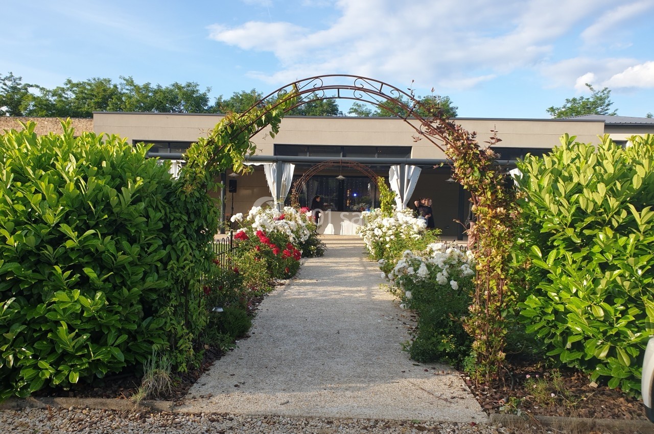 Allée bordée de buissons et de fleurs menant à une arche en fer forgé devant une terrasse avec rideaux blancs.