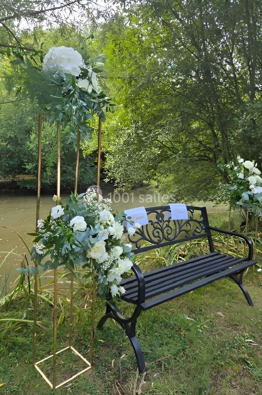 Banc en métal noir décoré de fleurs blanches et feuillages, placé près d'un cours d'eau entouré de verdure.