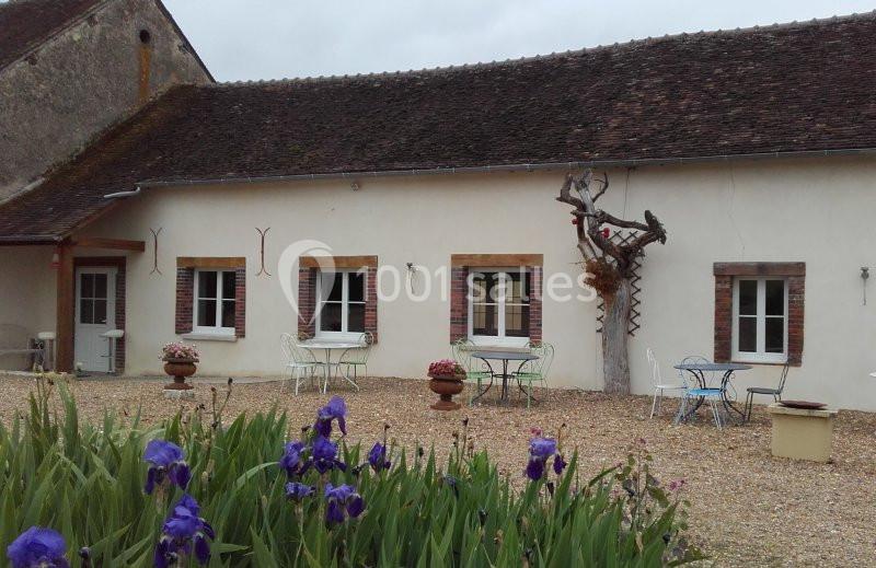 Façade d'une maison ancienne avec murs blancs, fenêtres encadrées de briques, tables extérieures et fleurs au premier plan.