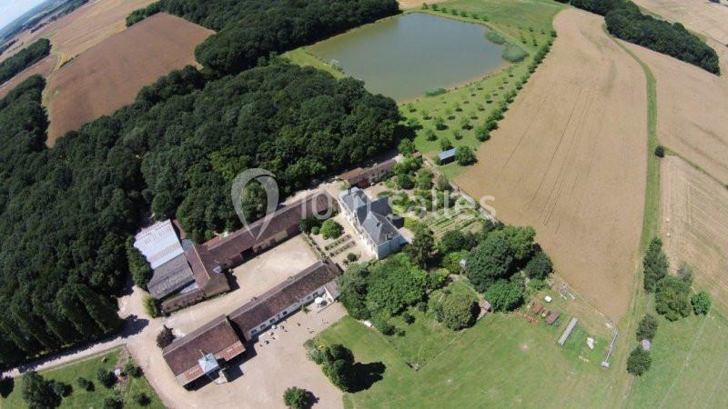 Vue aérienne d'un domaine rural avec bâtiments, forêt, champs cultivés et un étang bordé d'arbres.