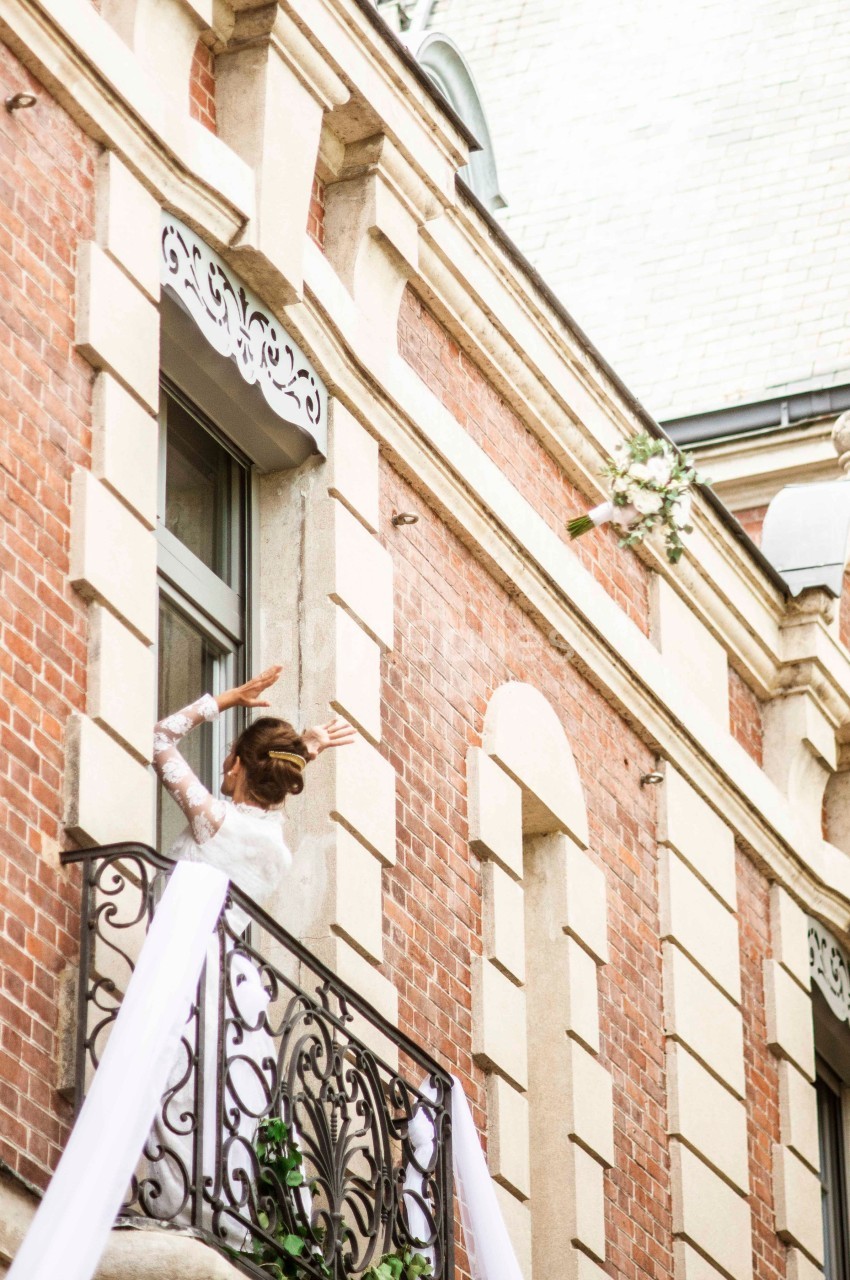 Femme en robe blanche saluant depuis un balcon décoré de tissu et de verdure sur une façade en briques.