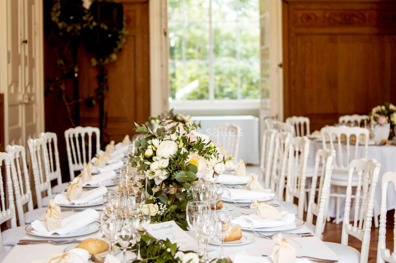 Salle décorée pour un mariage avec des tables rondes dressées, nappes blanches, fleurs et lumière naturelle.