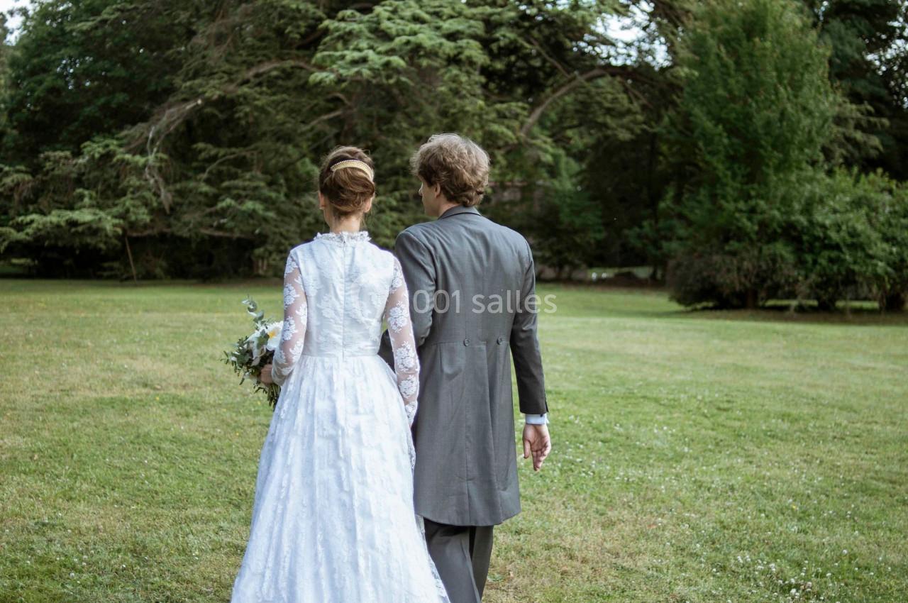 Un couple en tenue de mariage marche dans un parc verdoyant, vu de dos, sous un grand arbre.