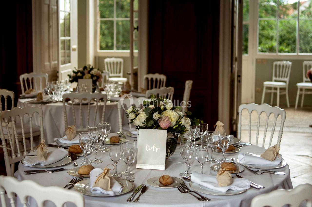 Salle de réception avec tables rondes dressées, nappes blanches, vaisselle élégante et bouquets de fleurs.