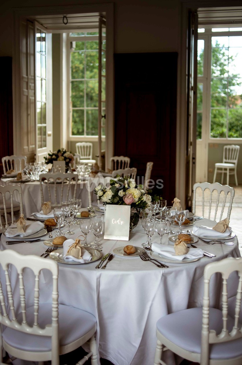 Salle de réception avec tables rondes dressées, nappes blanches, vaisselle élégante et bouquets de fleurs.