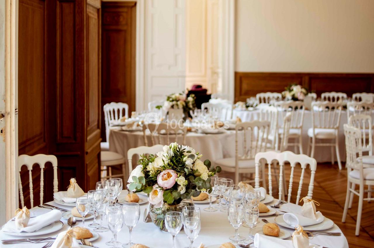Salle de réception élégante avec tables rondes dressées, nappes blanches, vaisselle raffinée et bouquets floraux.
