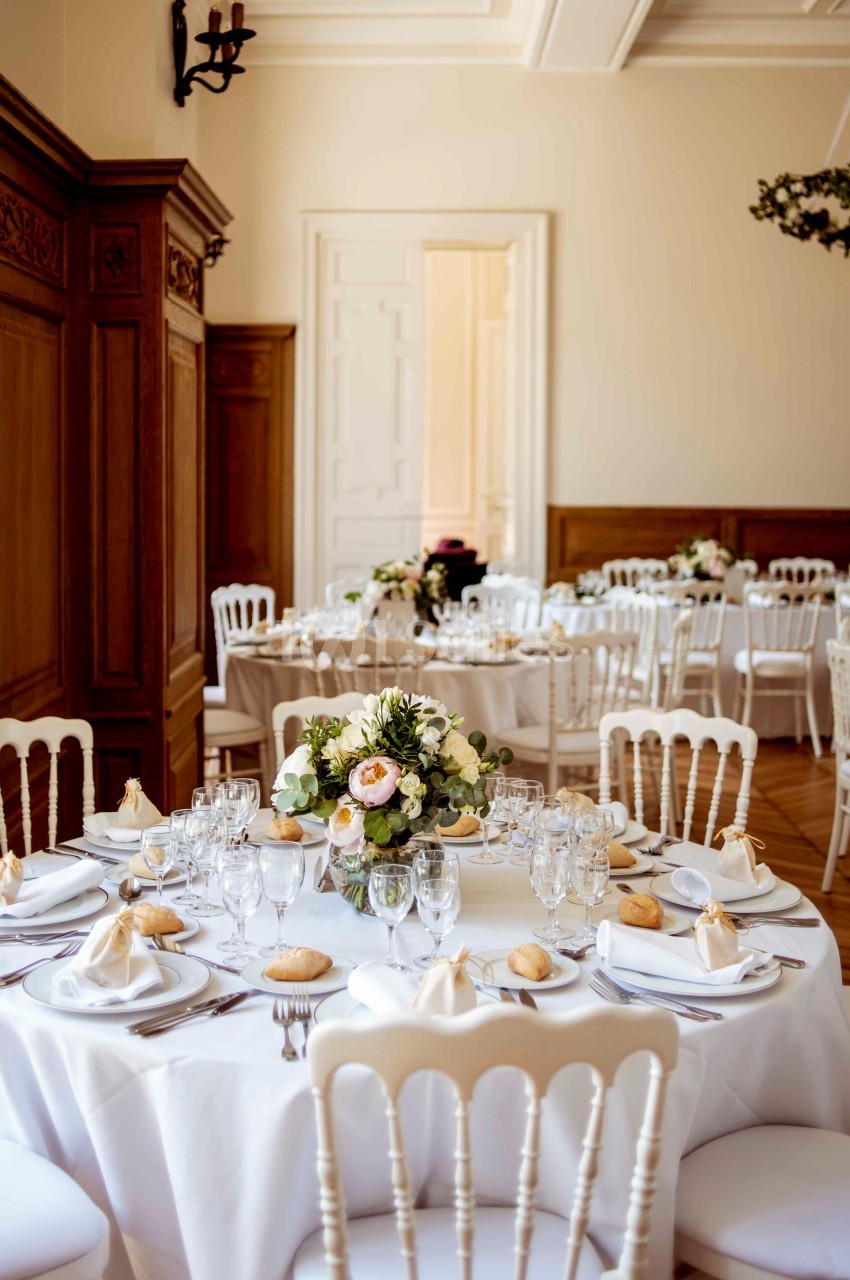 Salle de réception élégante avec tables rondes dressées, nappes blanches, vaisselle raffinée et bouquets floraux.