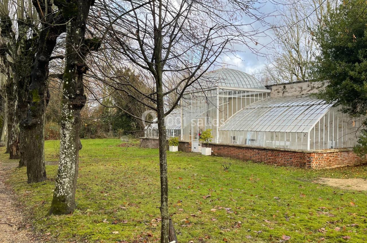 Serre en verre entourée d'arbres et d'une pelouse, située dans un jardin en hiver avec ciel nuageux.