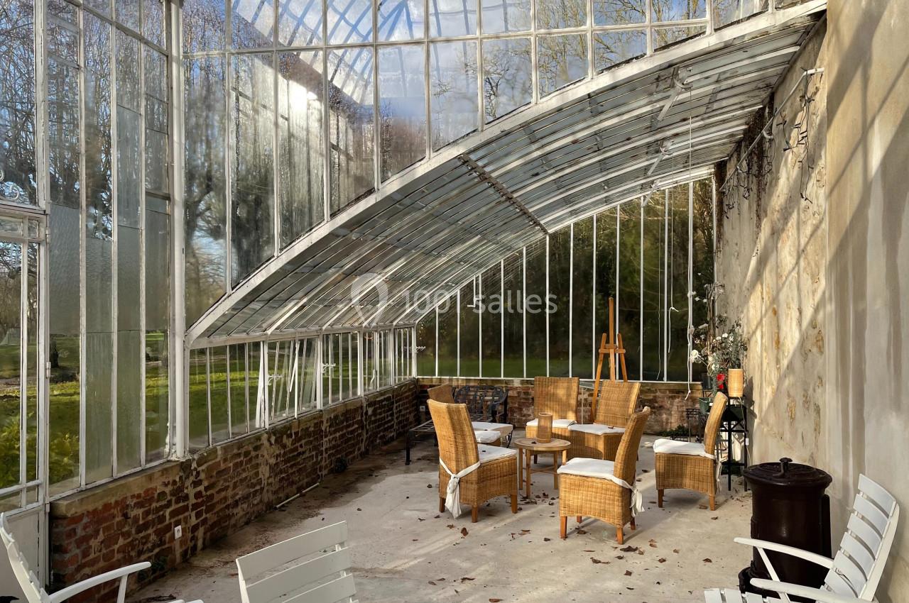 Vue intérieure d'une serre lumineuse avec des meubles en rotin et des chaises blanches, entourée de verdure extérieure.