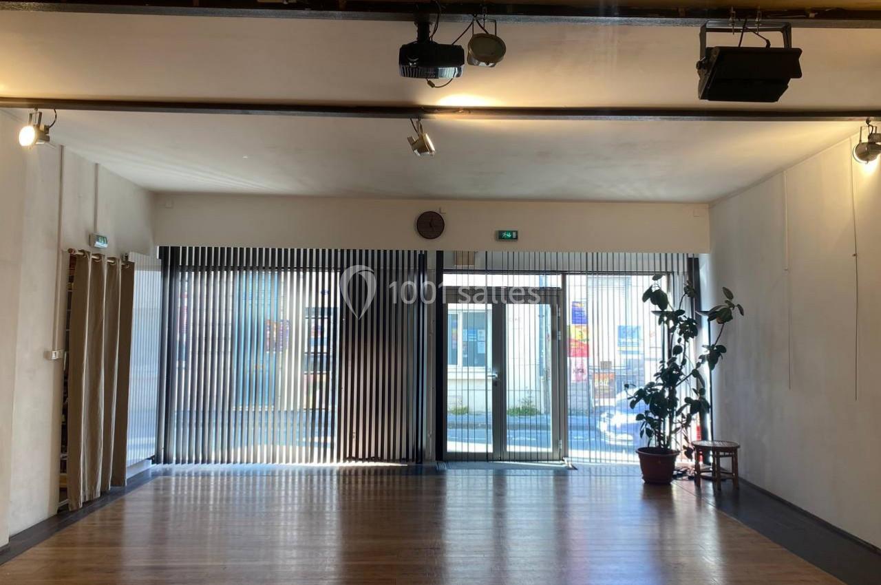 Salle lumineuse avec parquet, grande baie vitrée équipée de stores verticaux, et une plante en pot près de l'entrée.