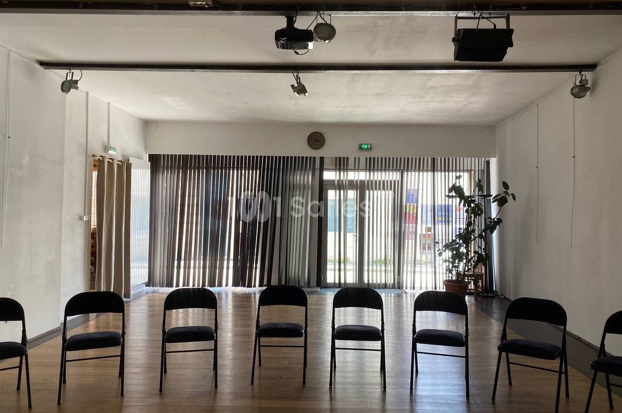 Salle lumineuse avec parquet, chaises noires alignées et grandes baies vitrées avec rideaux laissant passer la lumière.