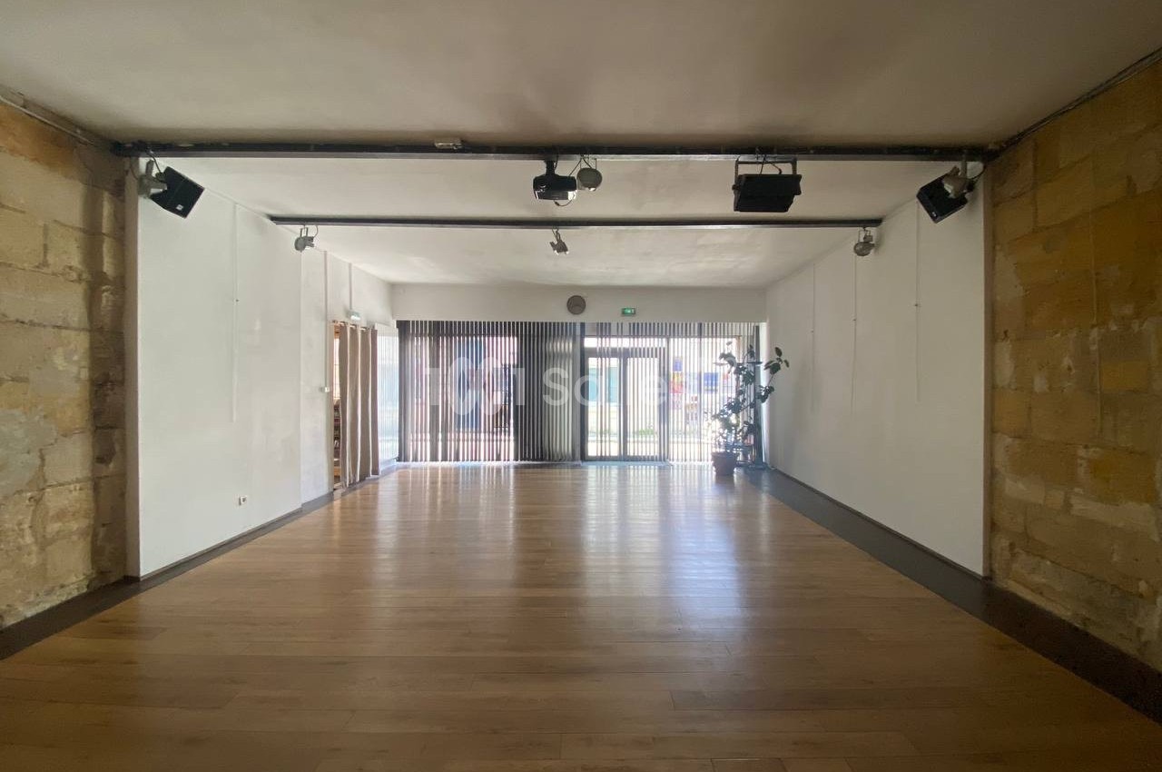 Salle vide avec parquet en bois clair, murs en pierre et en blanc, éclairage au plafond et grande baie vitrée au fond.
