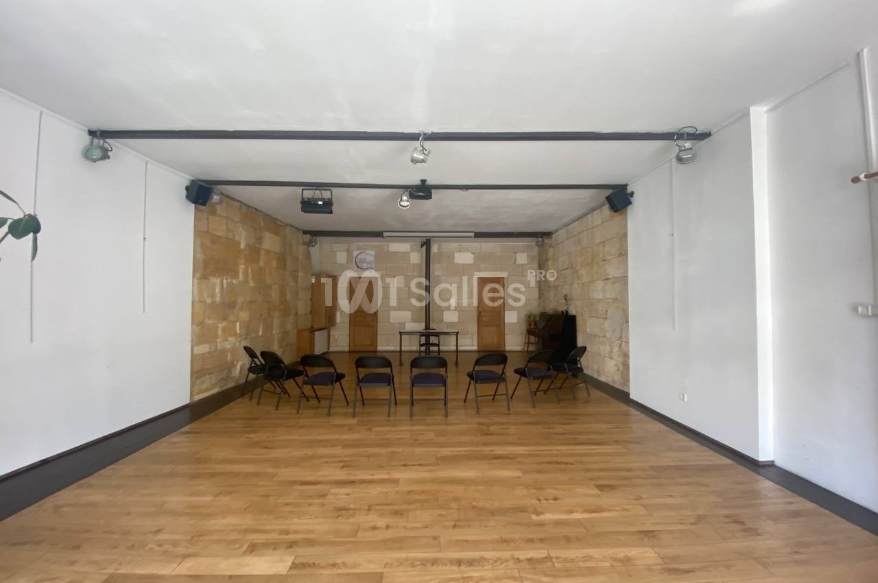 Salle lumineuse avec parquet en bois, murs en pierre et chaises disposées en cercle au centre.