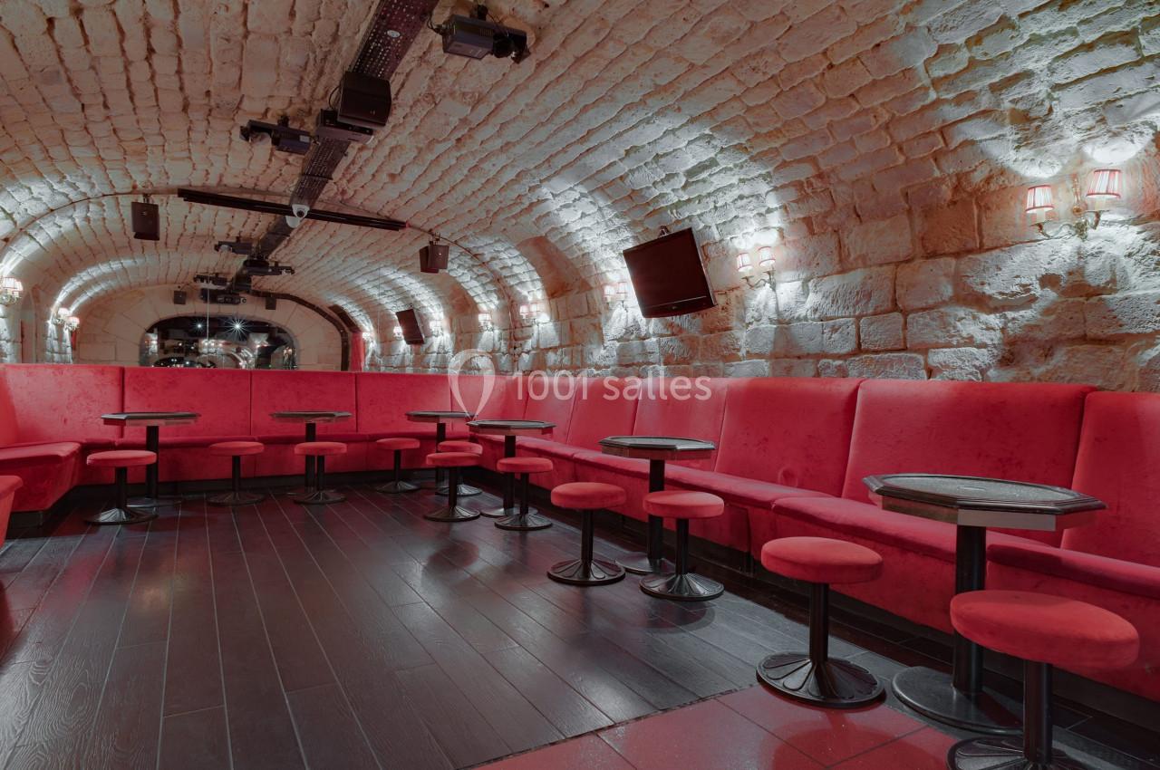 Salle voûtée en pierre avec banquettes rouges, tables basses et éclairage tamisé.